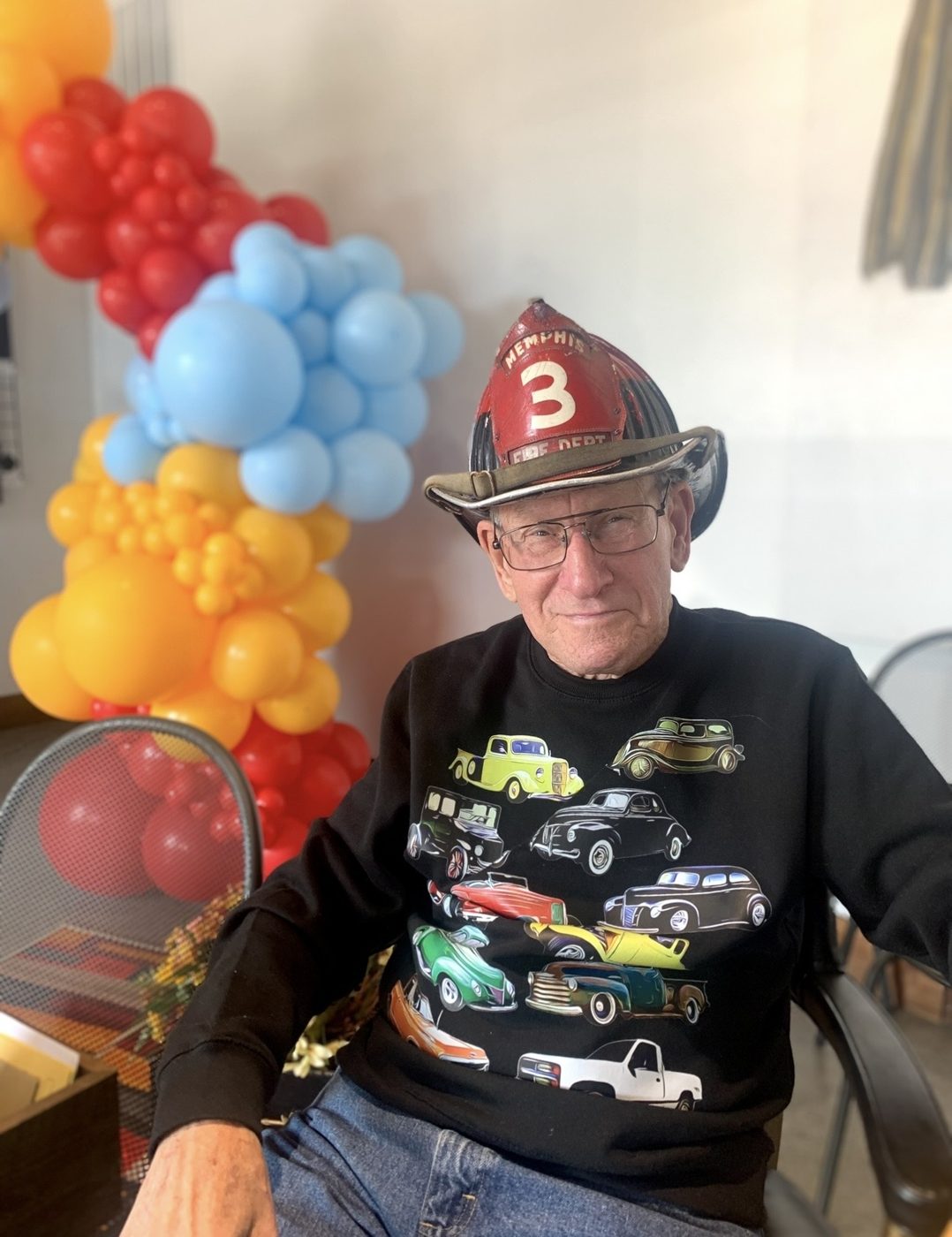 Buddy in a classic-car sweatshirt and a vintage Memphis Fire Department helmet beside red, yellow and blue balloons