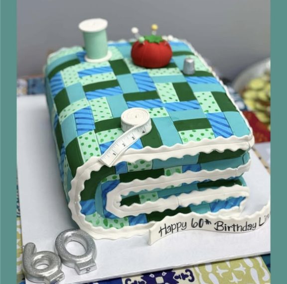 A cake shaped like a folded quilt with a spool, pincushion and tape measure on top