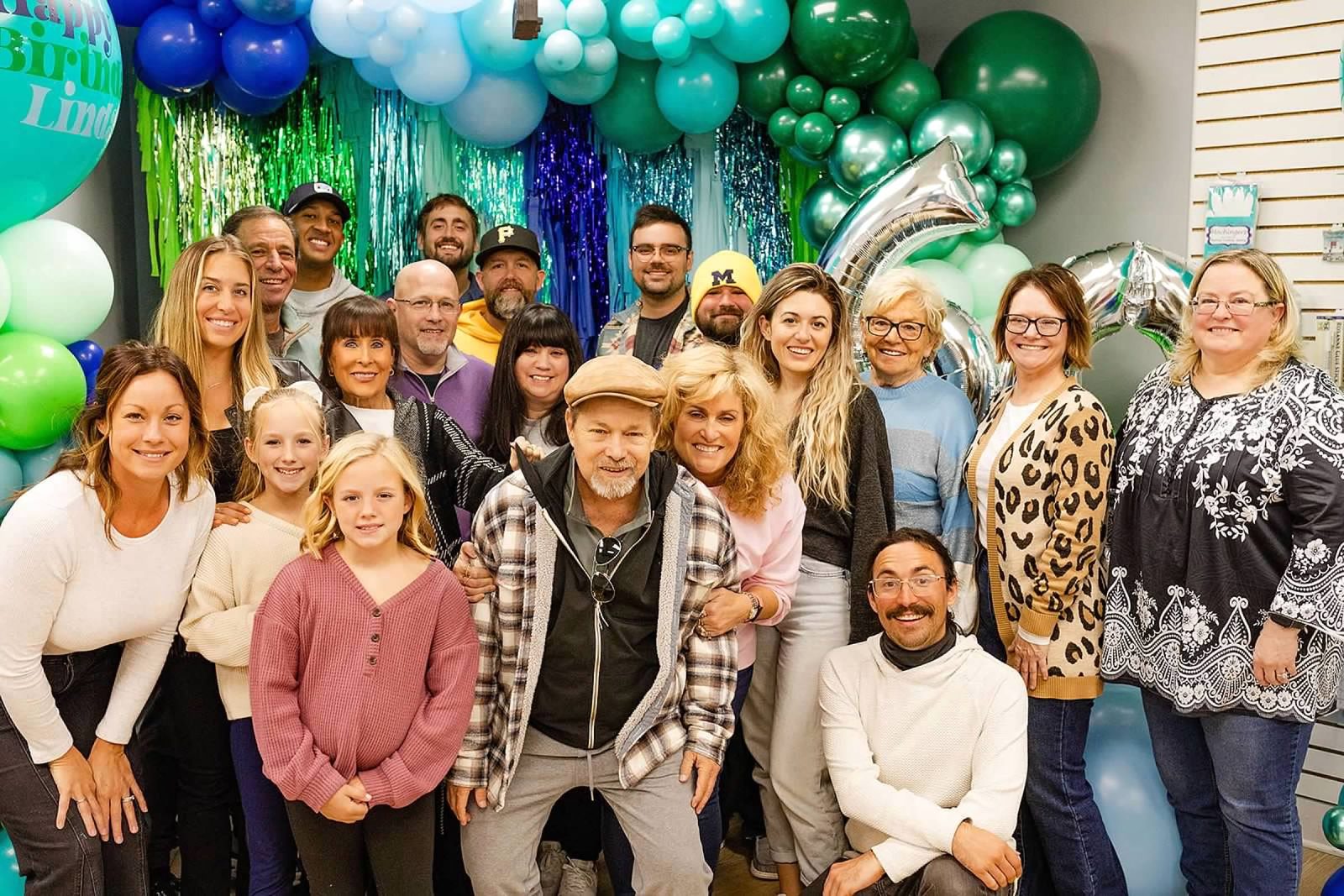 Family and friends gathered in front of a teal and green balloon backdrop at a surprise 60th birthday