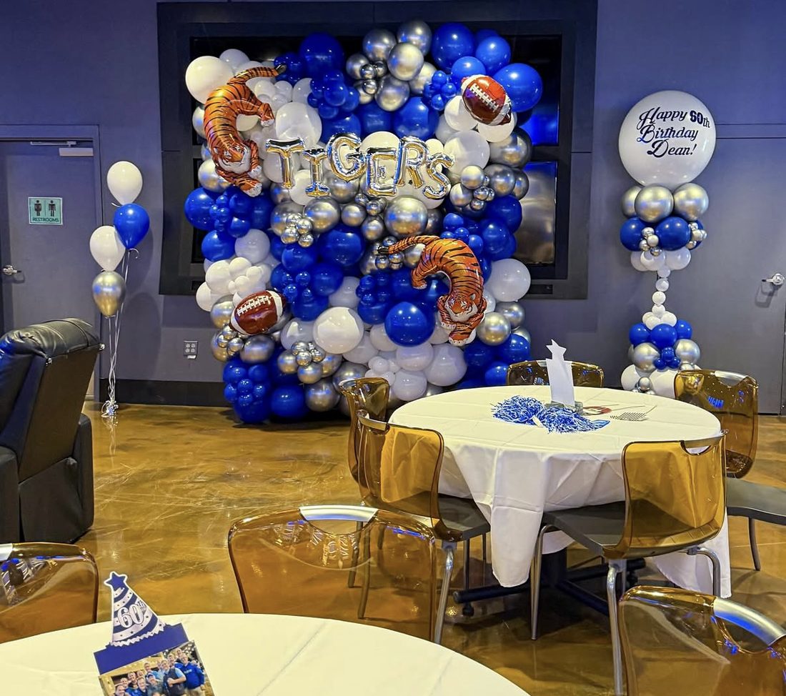A Memphis Tigers balloon wall in blue, white and silver with footballs and tiger foils, a birthday balloon column and pom-pom centerpieces