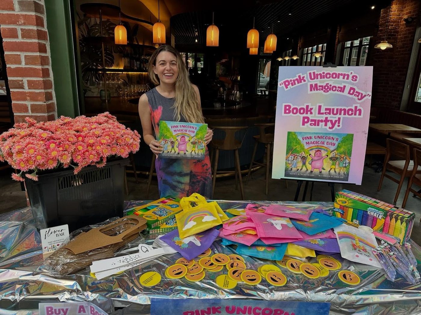 Nico holding Pink Unicorn's Magical Day beside a Book Launch Party sign, a flower bar and a table of rainbow kindness kits